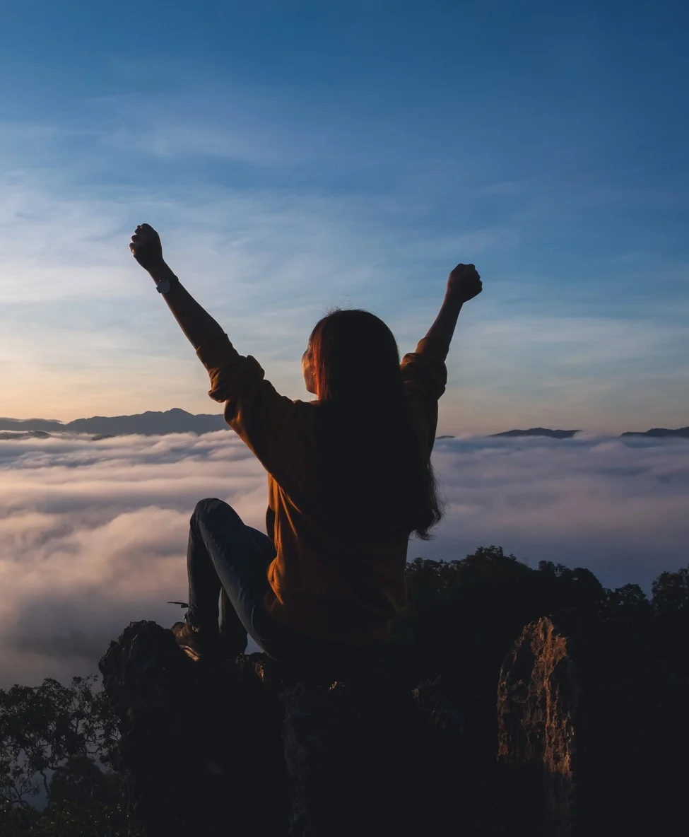 Woman sitting on a mountain ridge with arms raised watching the sunrise above the clouds, representing the calm and release somatic coaching and Qi Gong can bring to the nervous system